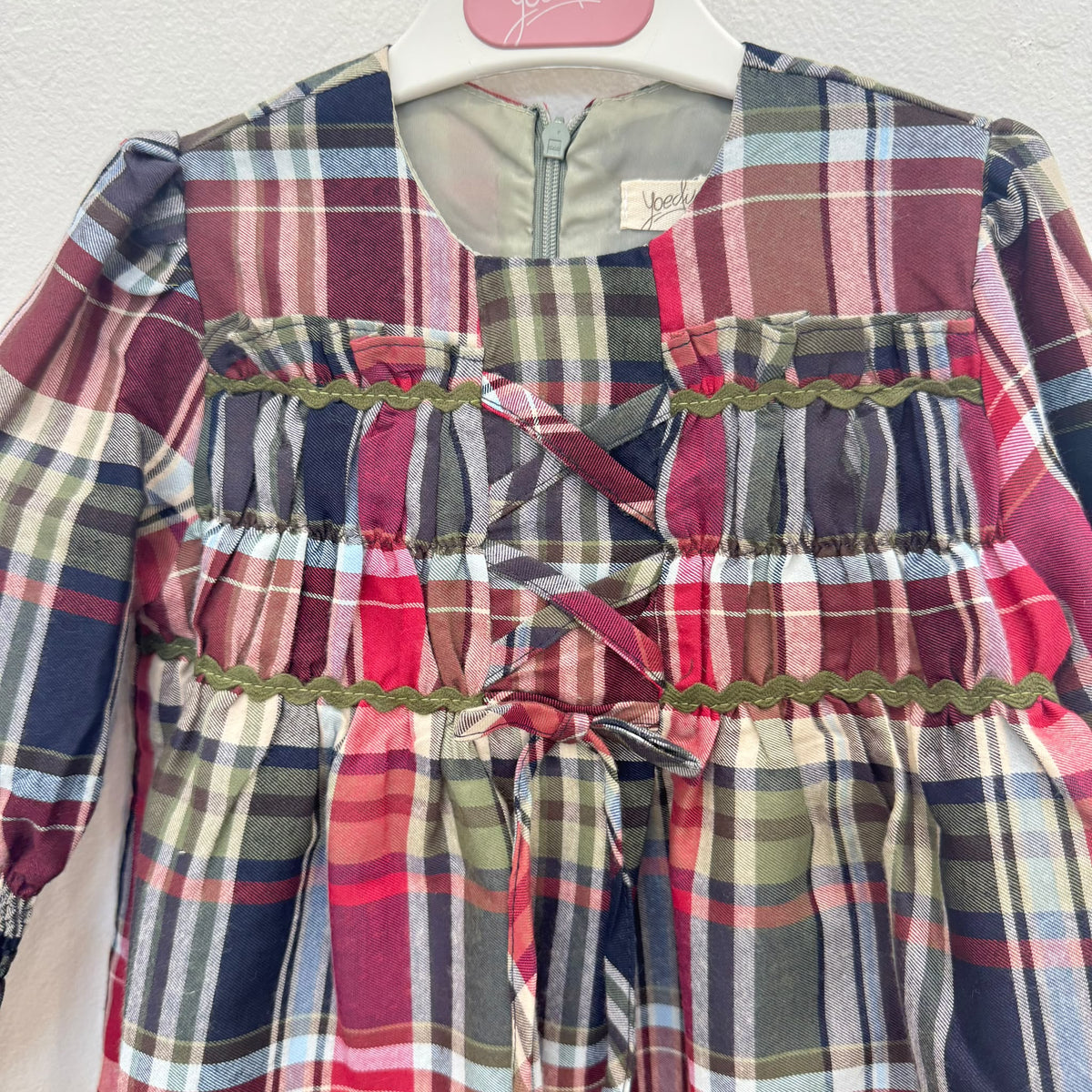 Abito bambina in fantasia tartan scozzese rosso, verde e blu, con maniche lunghe a volant, corpetto arricciato, passamaneria verde e fiocchi frontali.