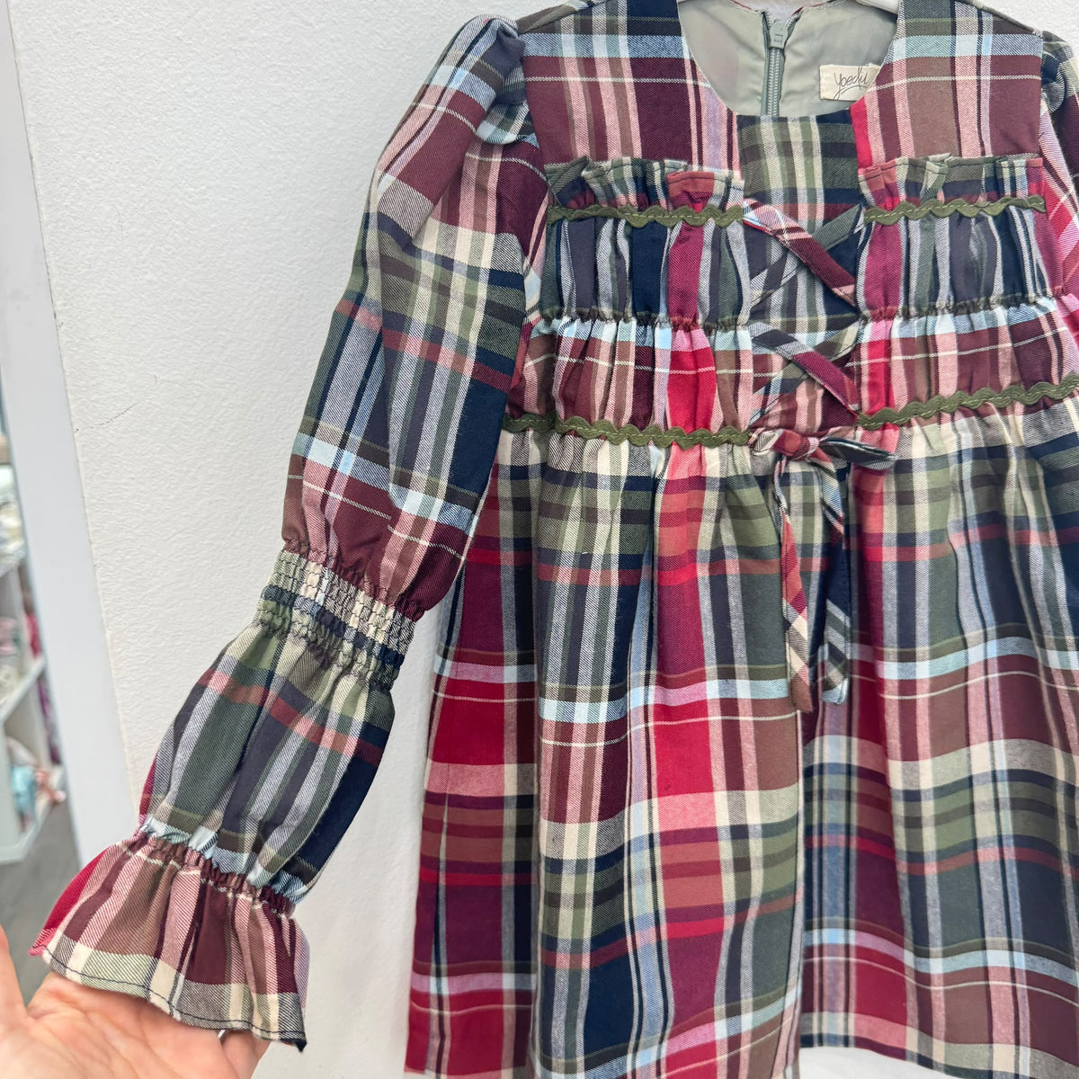 Abito bambina in fantasia tartan scozzese rosso, verde e blu, con maniche lunghe a volant, corpetto arricciato, passamaneria verde e fiocchi frontali.