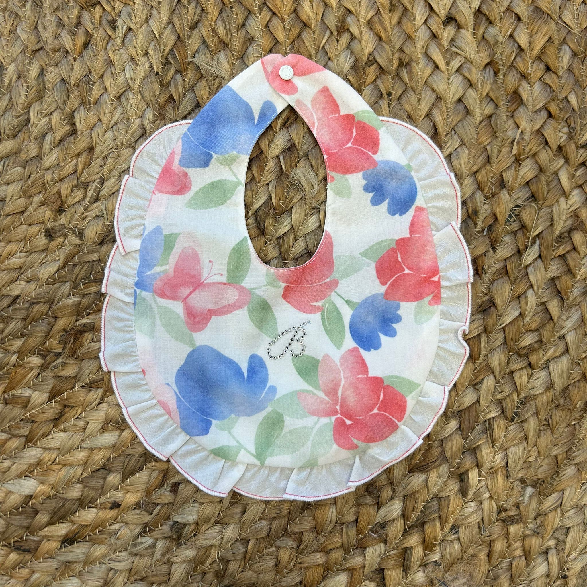 Bavaglino neonata in cotone con fiori e farfalle acquerello, balza arricciata e strass decorativi a forma di B.

