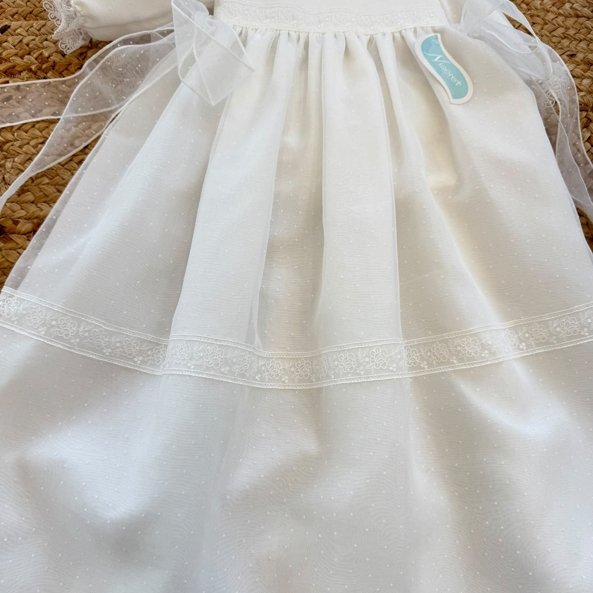 Dettaglio del tessuto a piccoli pois dell'abito battesimo neonata, con ricami floreali bianchi e bordo in pizzo smerlato.