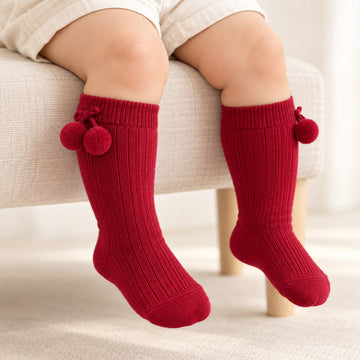 Calzettoni neonato unisex rosso bordeaux a costine con morbidi pompon laterali. Eleganti e raffinati, perfetti per pagliaccetti e completini nascita.