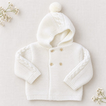 Cappottino battesimo bimbo bianco latte in maglia con cappuccio e pompon, trecce decorative sulle maniche e chiusura doppiopetto con bottoni chiari.
