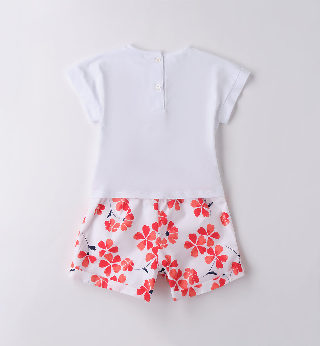 Outfit bambina estivo con t-shirt bianca a cuore floreale e pantaloncini a fiori rossi e blu, ideale per cerimonie informali e eventi primaverili.