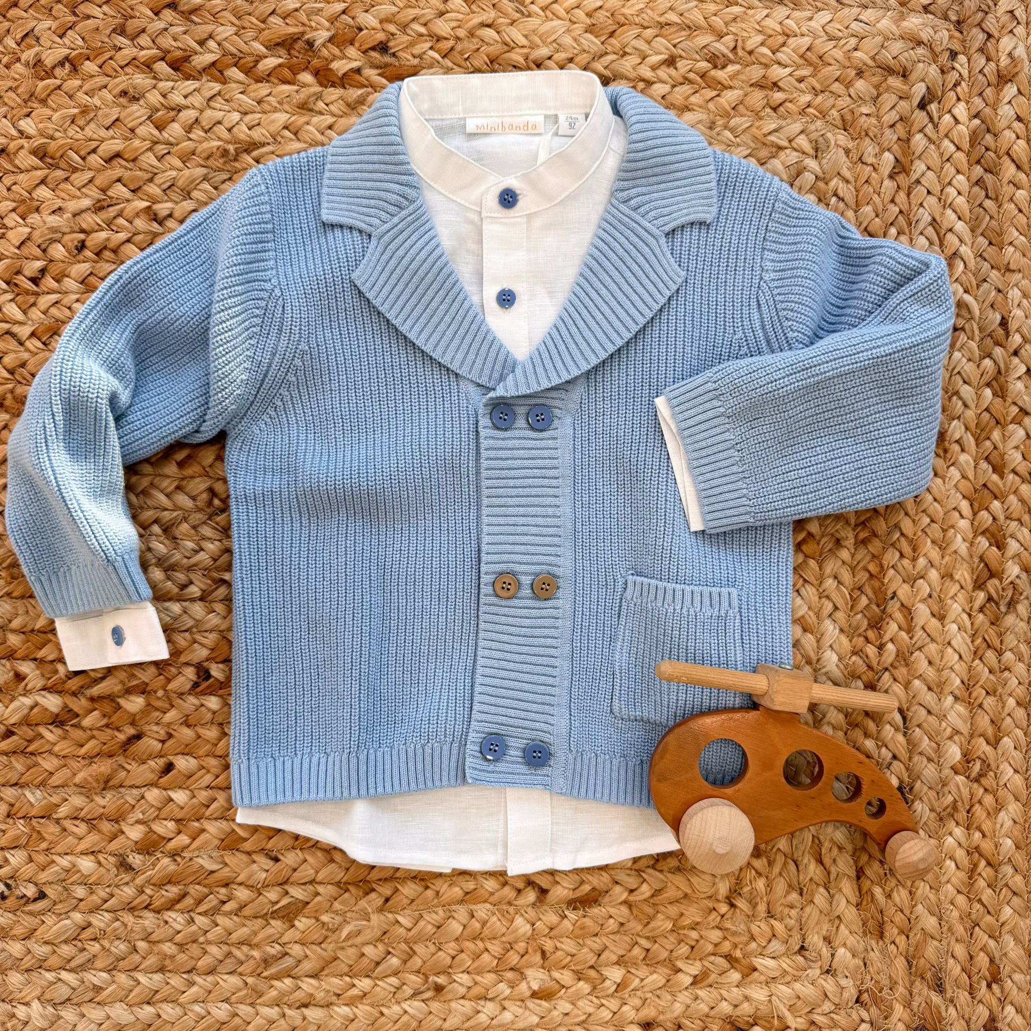 Completo neonato con camicina bianca e cardigan blu polvere in maglia, accanto a un elicottero in legno.