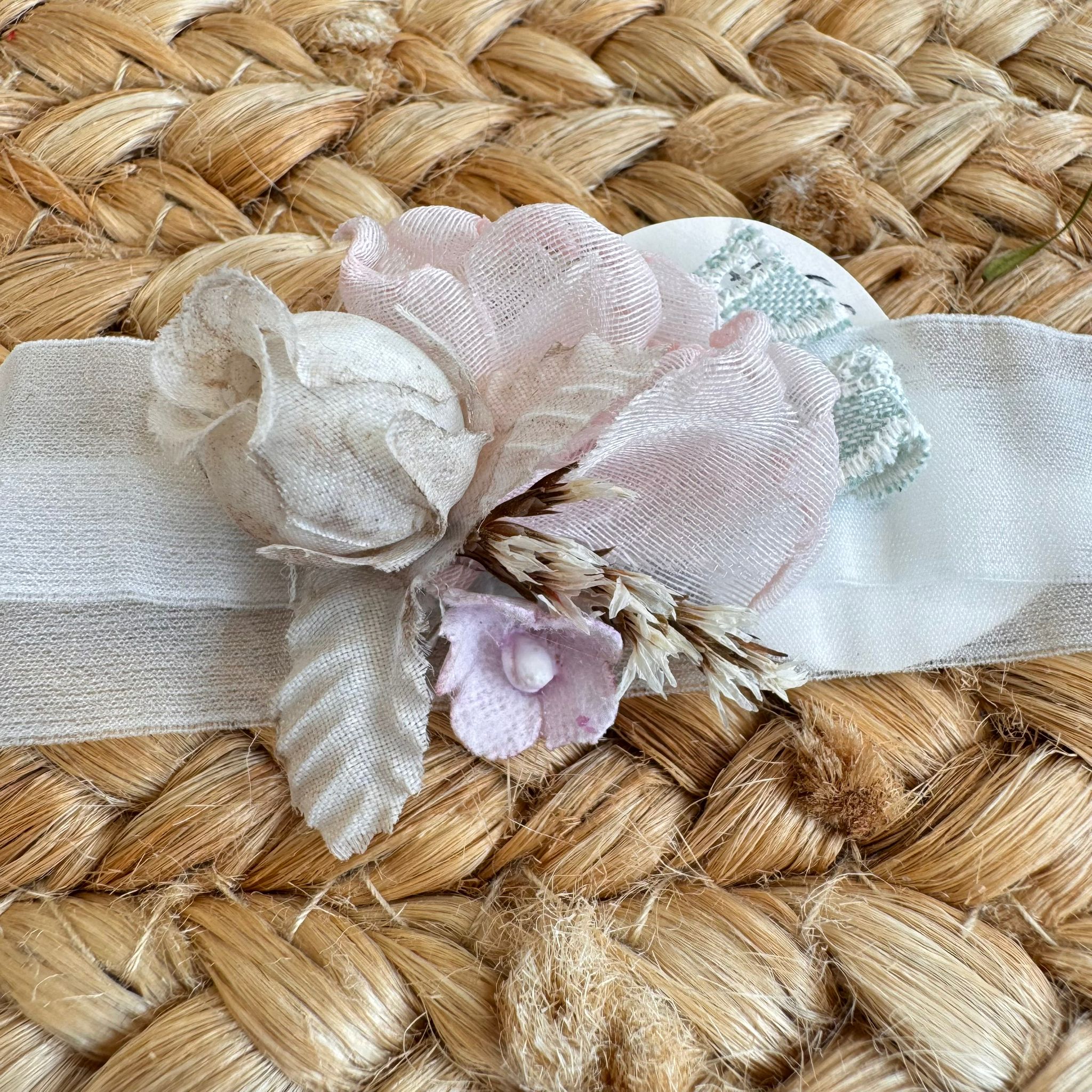 Fascia neonata per battesimo con fiori rosa cipria e spighe naturali su sfondo intrecciato