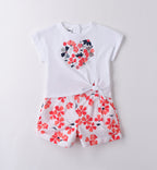 Outfit bambina estivo con t-shirt bianca a cuore floreale e pantaloncini a fiori rossi e blu, ideale per cerimonie informali e eventi primaverili.