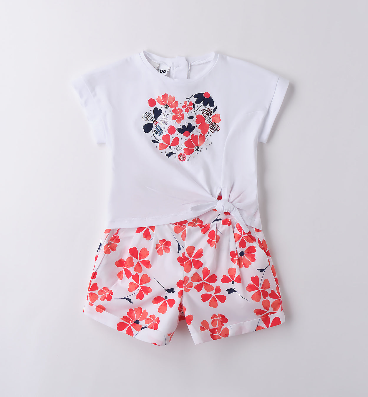 Outfit bambina estivo con t-shirt bianca a cuore floreale e pantaloncini a fiori rossi e blu, ideale per cerimonie informali e eventi primaverili.