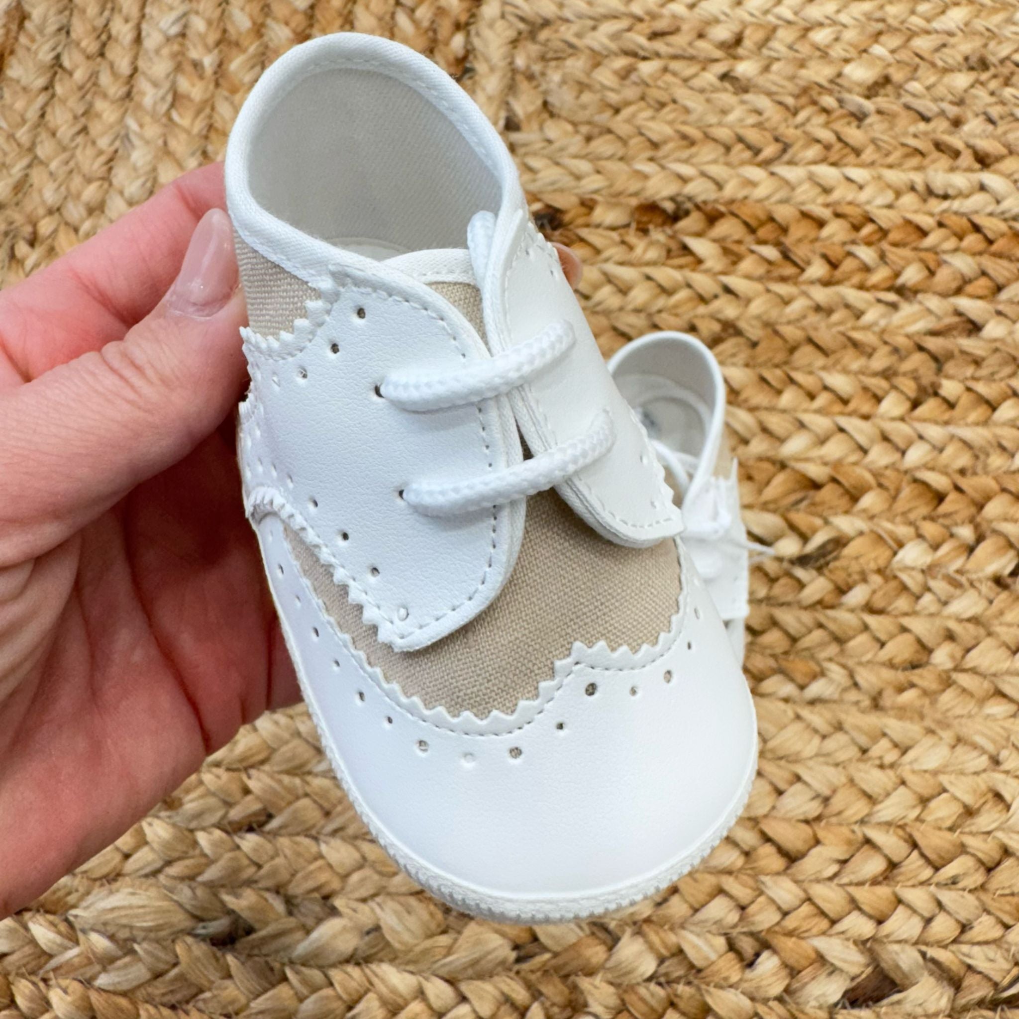 Scarpe culla dallo stile classico con tomaia in pelle e tessuto beige, impreziosite da dettagli traforati e chiusura a lacci.