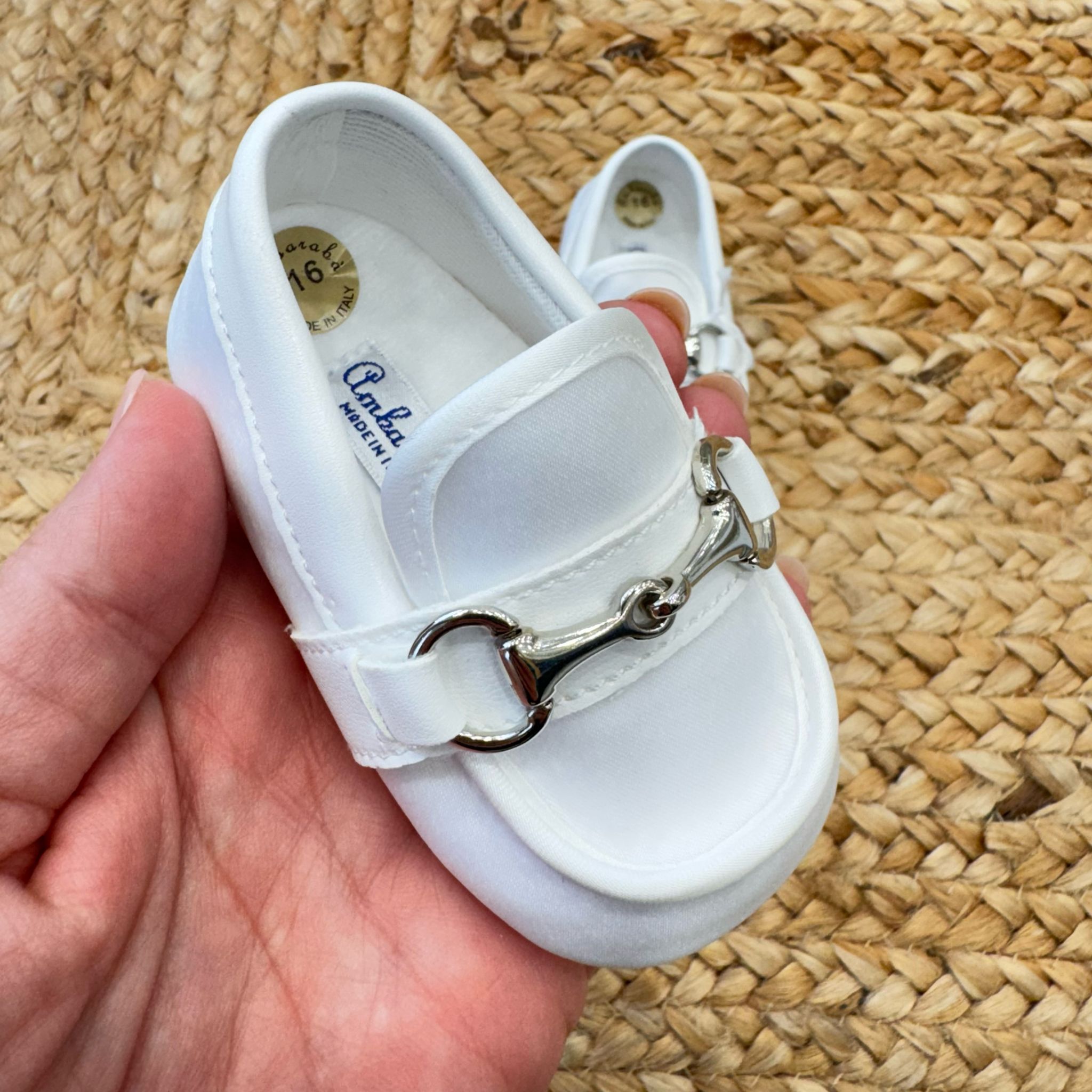 Scarpe culla stile mocassino in raso bianco con dettaglio metallico frontale, ideali per battesimo e cerimonia.