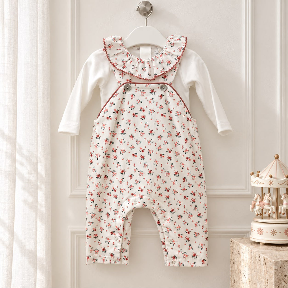 Tutina neonata in cotone con salopette bianca a piccoli fiori rossi e rosa, maglia bianca a maniche lunghe, ampio colletto volant floreale, bottoni sulle bretelle e profili color rosso mattone.