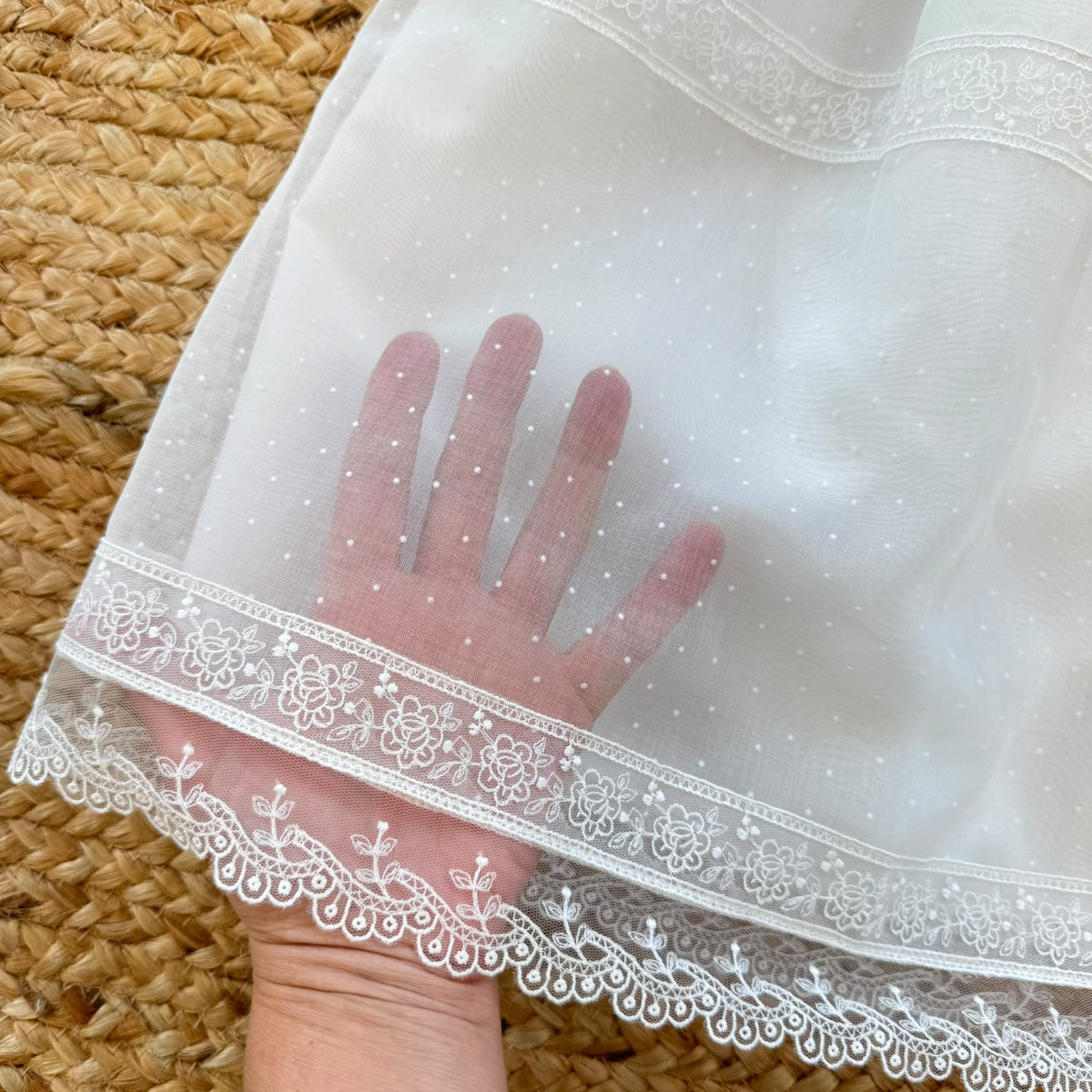 Dettaglio del tessuto a piccoli pois dell'abito battesimo neonata, con ricami floreali bianchi e bordo in pizzo smerlato.