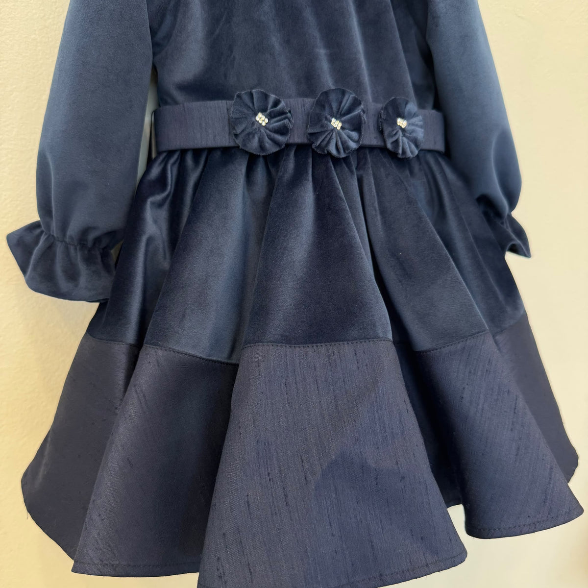 Abito elegante bimba blu in velluto con colletto arrotondato, maniche lunghe e tre fiori decorativi sul punto vita.